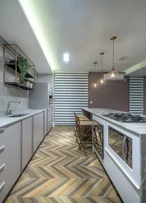 Cozinha com um piso em paginação espinha de peixe com cores diferentes em tons cinzas e marrom. Lampadas douradas, decoração de plantas e balção com bancos de madeira.