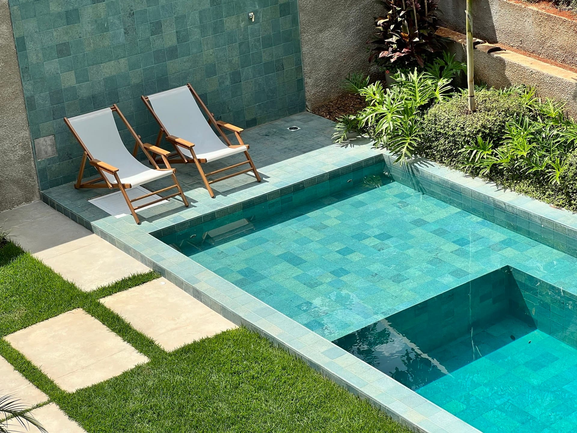 Piscina encostada em muro. Há plantas e espreguiçadeiras ao redor dela