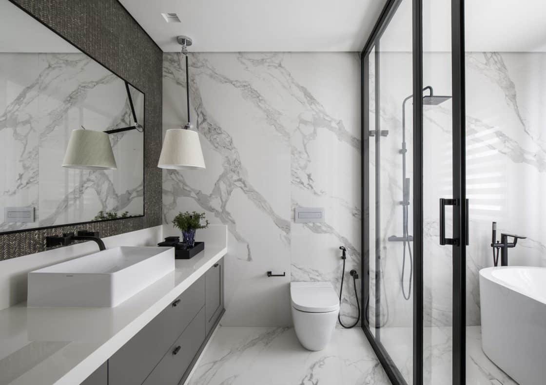 Box com dois chuveiros permite ao casal tomar banho ao mesmo tempo com conforto. Neste projeto, o dourado aparece nos metais, no revestimento brilhante e até mesmo nos veios do porcelanato que reproduz o mármore.