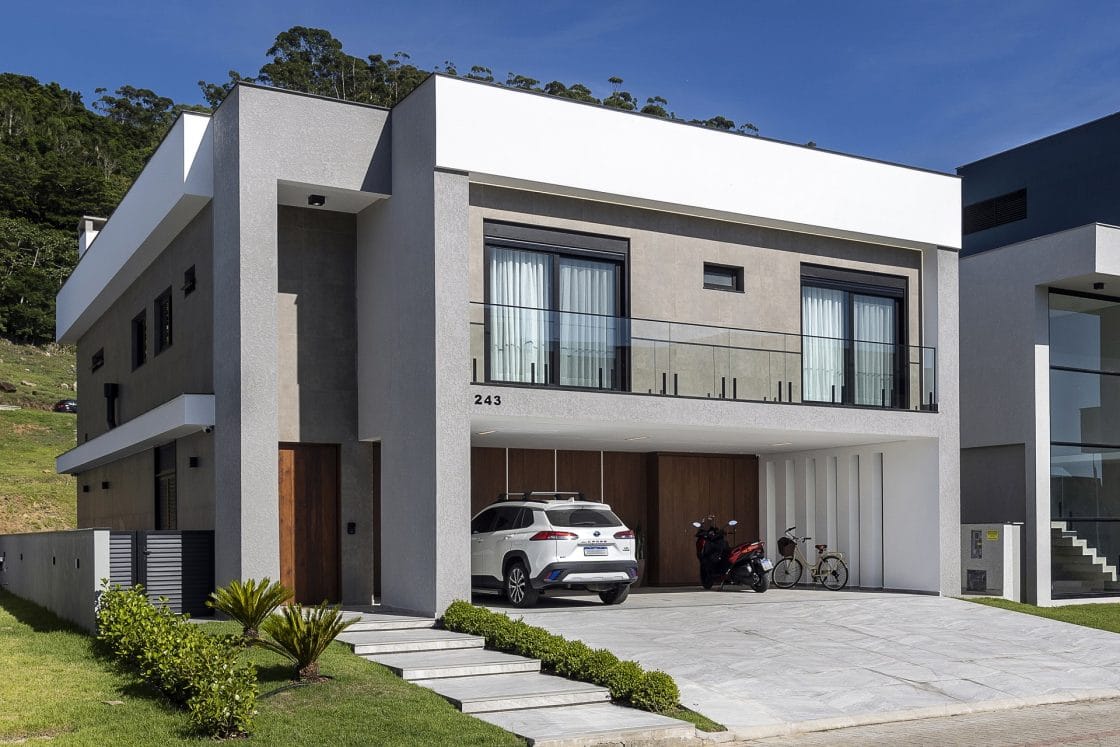 Casa com linhas retas e cores neutras. Na garagem há um carro, uma moto e uma bicicleta (Projeto: Dalma Meneses Arquitetura)