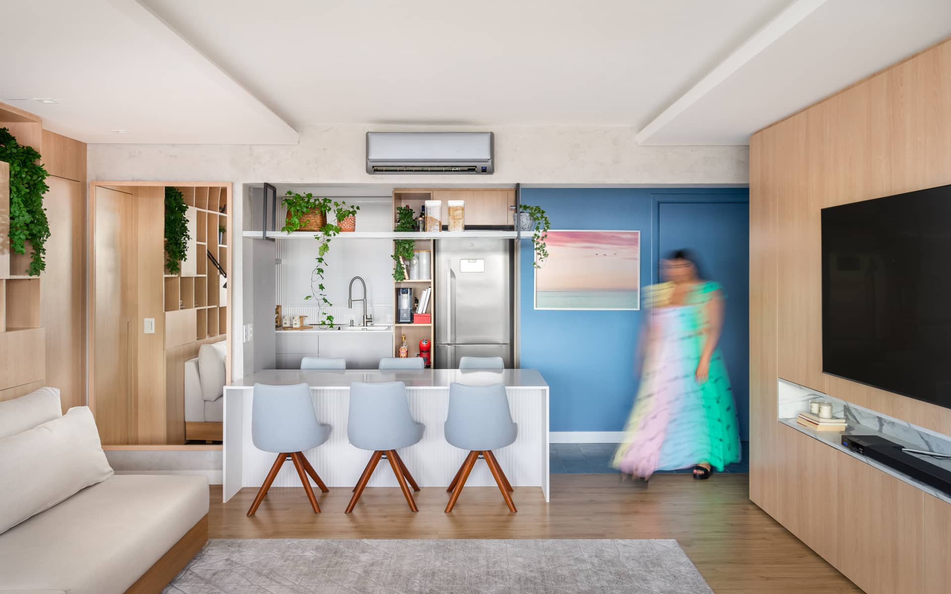 Sala em tons claros, com parede azul e porta pintada na mesma cor. Há banquetas, estante, painel em madeira com TV e sofá.