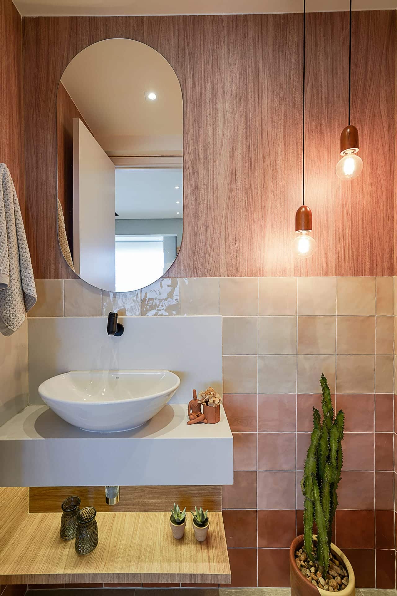 Banheiro com revestimento em madeira acima. Na parte de baixo, um degradê de revestimentos em diferentes tons alaranjados. Há luminárias, espelho e bancada clara.