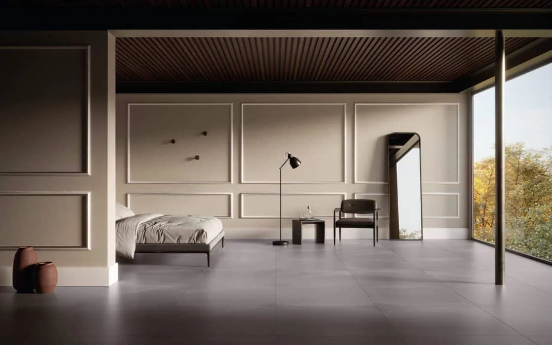 Quarto em tom acinzentado, com molduras de boiserie na parede. Há a cama, uma luminária, cadeira e espelho.